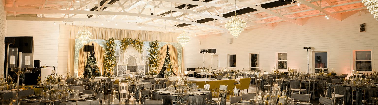 The grand ballroom set for a wedding reception: exposed white trusses, crystal chandeliers, a floral stage and dozens of dressed tables